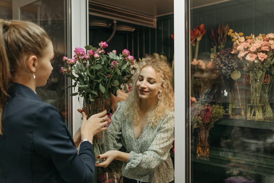A woman with curly blonde hair, dressed in a light patterned blouse, is receiving a bouquet of pink roses from a florist at a flower shop. The florist, dressed in a navy blazer, hands over the arrange