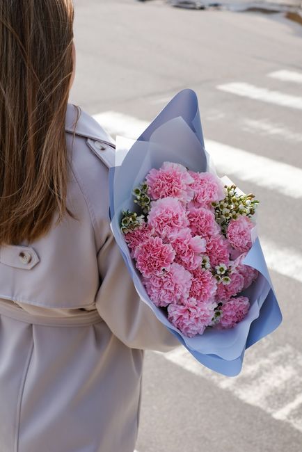 A person dressed in a light-colored coat is holding a floral gift wrapped in blue paper, featuring a bouquet of pink carnations and small white daisies with green foliage. The arrangement appears fres