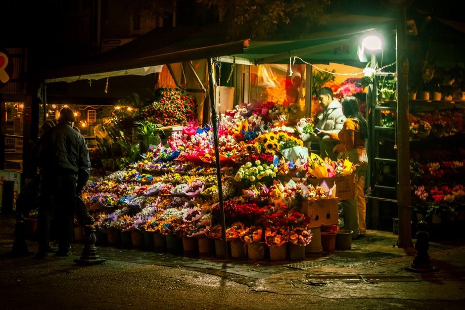 At a bustling flower stall during the evening, a wide array of fresh flowers including vibrant sunflowers, roses, lilies, and chrysanthemums are displayed in buckets and wrapped bouquets under bright 