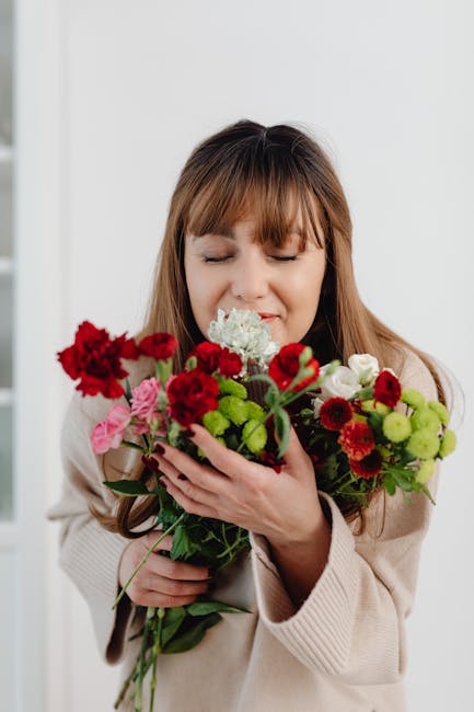A woman with light brown hair and bangs, dressed in a beige coat, is holding a vibrant floral bouquet close to her face with her eyes gently closed, appearing to enjoy the fragrance. The bouquet featu