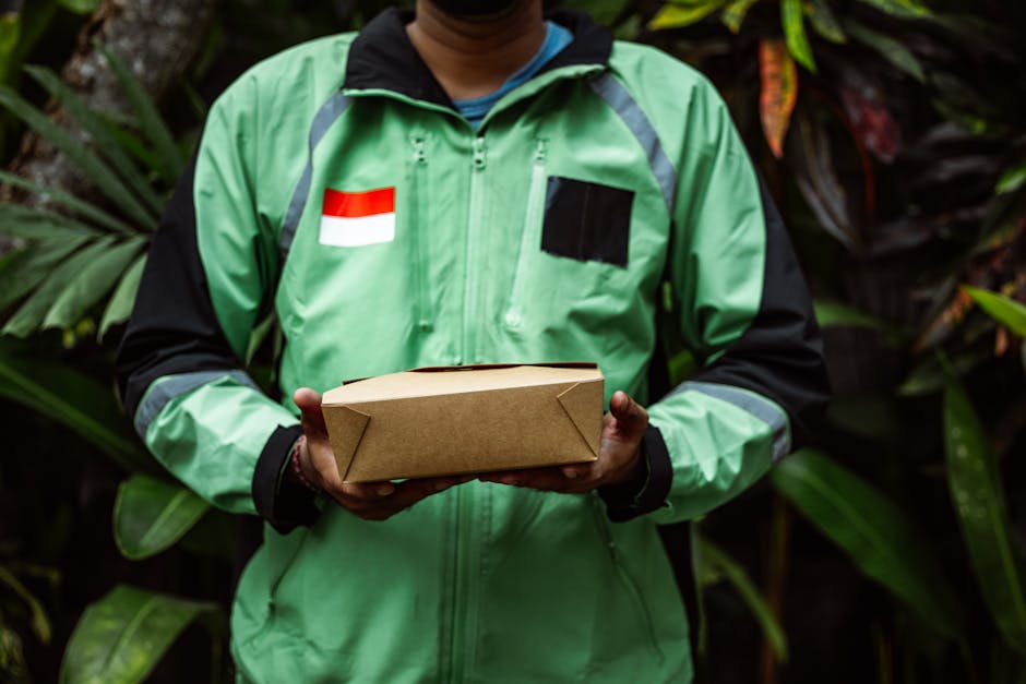 A person wearing a bright green delivery jacket with black sleeves, featuring a red and white flag patch and a black rectangular patch, stands amidst lush green foliage. They hold a rectangular brown 