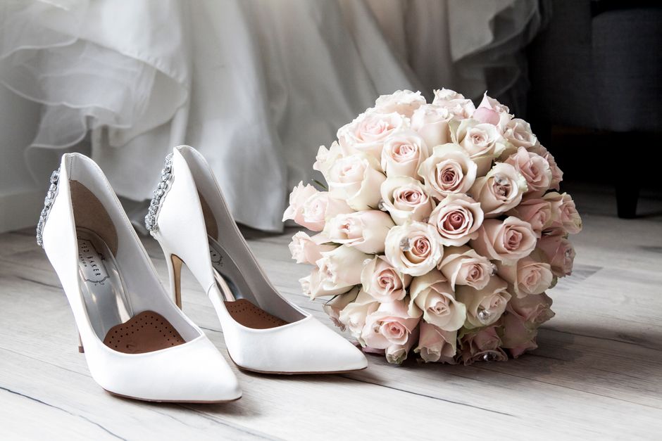 A pair of elegant white high-heeled shoes with pointed toes and decorative embellishments on the back, positioned on a light wooden floor. Next to the shoes is a large, rounded bouquet of fresh pale p
