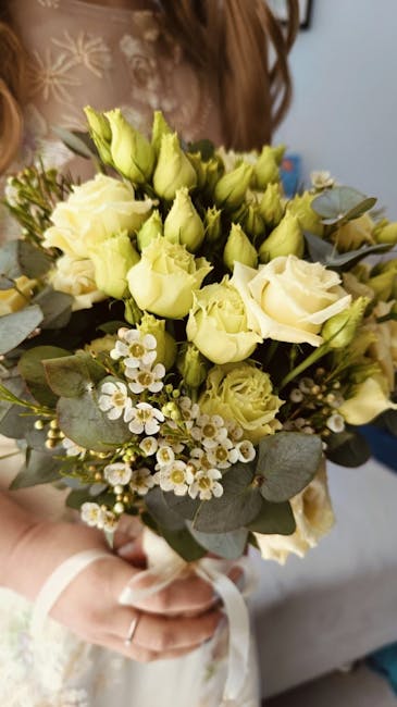 A person holding a bouquet of fresh cream and pale yellow roses, accented with small white blossoms and green eucalyptus leaves. The roses are in full bloom with soft, velvety petals, arranged in a ro